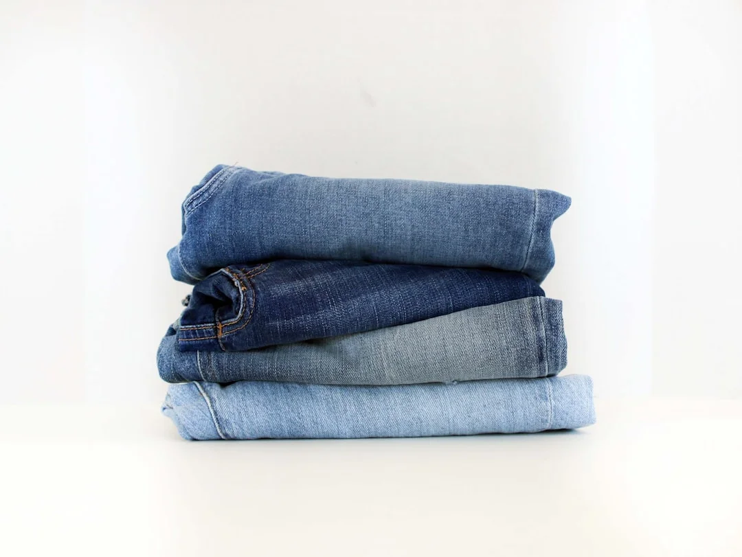 A woman standing in natural light beside an open wardrobe rail, holding a folded pair of dark indigo jeans and comparing them to a lighter wash pair draped over her arm